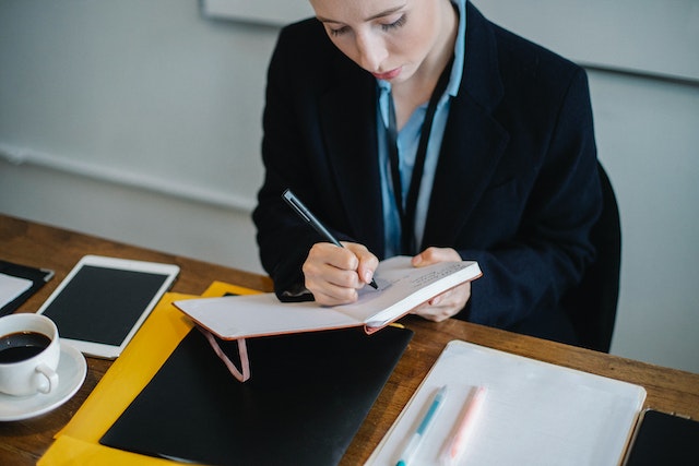 person in black blazer writing down a note in their notebook while sitting at their dark wood desk