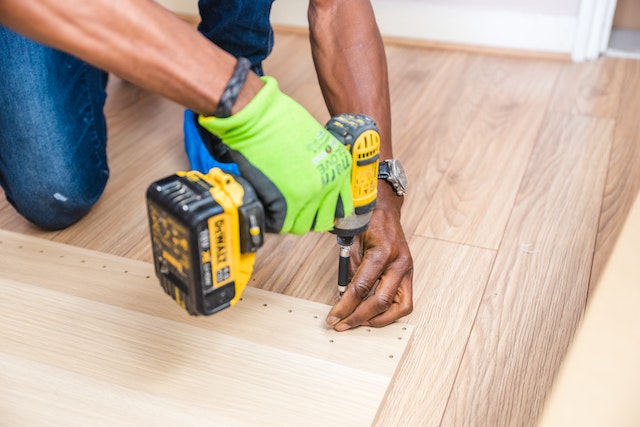 person in green work gloves using an electric drill on a piece of wood